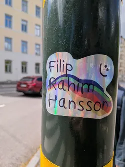 A custom-shaped sticker with an irregular, cloud-like outline and a thin white border. The background is highly iridescent and holographic, reflecting a spectrum of colors including pink, purple, blue, green, and yellow. Black, handwritten-style text is prominently displayed across the sticker, reading 'Filip' on the top line, 'Rahim' on the middle line, and 'Hansson' on the bottom line. A simple, hand-drawn black smiley face (two dots for eyes, a curved line for a mouth) is located in the top right corner. A subtle rainbow arc, with visible bands of blue, green, yellow, orange, and red, is integrated into the design, appearing behind and through the text.