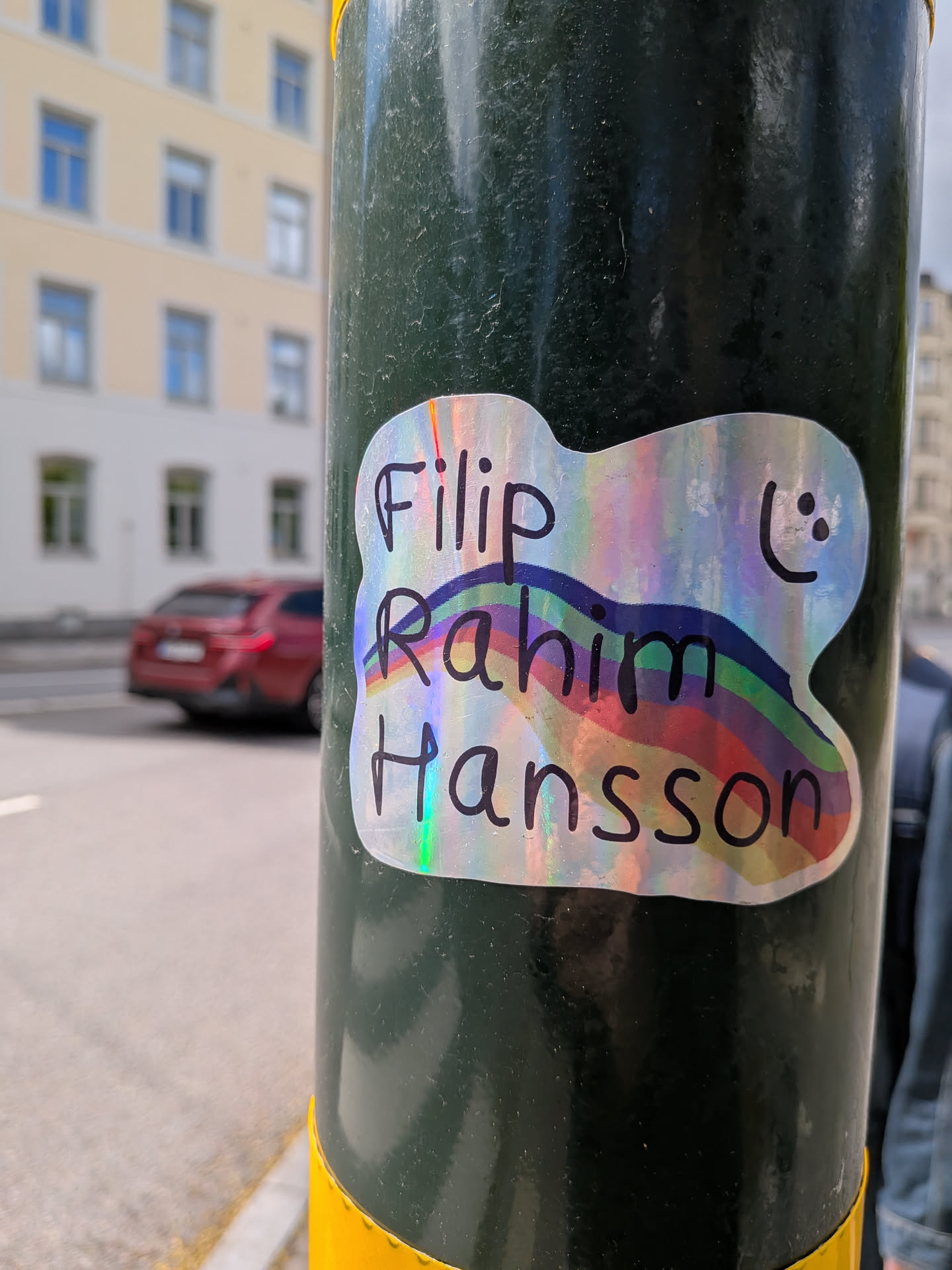 A custom-shaped sticker with an irregular, cloud-like outline and a thin white border. The background is highly iridescent and holographic, reflecting a spectrum of colors including pink, purple, blue, green, and yellow. Black, handwritten-style text is prominently displayed across the sticker, reading 'Filip' on the top line, 'Rahim' on the middle line, and 'Hansson' on the bottom line. A simple, hand-drawn black smiley face (two dots for eyes, a curved line for a mouth) is located in the top right corner. A subtle rainbow arc, with visible bands of blue, green, yellow, orange, and red, is integrated into the design, appearing behind and through the text.