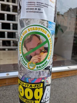 Street sticker A circular sticker featuring a man eating a burger, overlaid with a green 'no' symbol (a circle with a diagonal line). The man has light brown hair and is seen from the chest up, with only part of his face visible behind the burger. The outer ring of the sticker is green, with yellow text on the top half reading 'SÖDER IST EIN STADTTEIL IN STOCKHOLM' and green text on the bottom half reading 'TYKSA BAJARE'. At the very bottom, centered, is a small emblem resembling a flag or crest with green and white stripes, possibly the Hammarby IF (HIF) logo, surrounded by a golden wreath. The sticker has a white inner border separating the central image from the text ring.