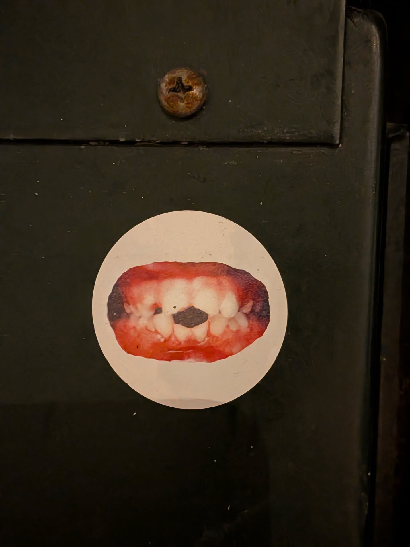 The sticker is circular with a white background. At its center is a photographic image of an open human mouth, focusing on the teeth and gums. The gums are a vibrant reddish-pink, while the teeth are off-white with some yellowing. A prominent, dark, irregular gap is visible between the two upper front teeth, creating a central void. The overall image has a slightly unsettling or raw aesthetic, with visible details of the oral cavity.