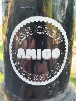 Black and white circular sticker with the text "ESH CREW" at the top, "AMIGO" in the center, and "STOCKHOLM" at the bottom. The text has a 3D effect. The sticker is on a dark surface.