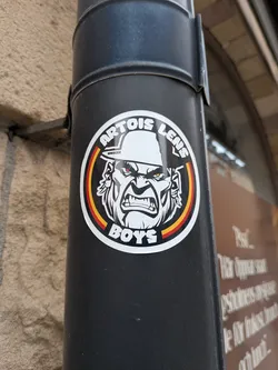 Street sticker A circular sticker featuring a stylized, aggressive male face with a furrowed brow, bared teeth, and glowing red/yellow eyes, wearing a helmet. The face is rendered in black and white with grey shading. An inner ring around the face displays vertical stripes of red, yellow, and black. The text "ARTOIS LENS" is arched at the top, and "BOYS" is arched at the bottom, both in bold black capital letters. A small Roman numeral "HHXXII" is visible on the helmet below "ARTOIS LENS". The sticker has a white background and a thick black outer border.