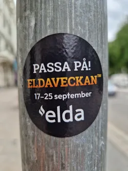 Street sticker Round black and orange sticker on a metal pole.  The text says "PASSA PÅ! ELDAVECKAN" with the dates "17-25 september" below it. There is also a logo that says "elda".