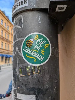 Street sticker A circular sticker with a dark teal-green background and a white border. On the left, a stylized golden beer mug with white foam is depicted. To its right, the text 'Öl på' is written in a white, elegant script font, positioned above 'SÖDERMALM' in a bold, white, sans-serif, all-caps font. Yellow stylized 'splashes' or 'rays' emanate from the text, and a yellow decorative swirl underlines 'SÖDERMALM'. The overall design is clean, cheerful, and inviting.