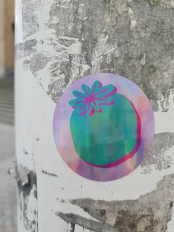 Close-up of a circular sticker affixed to a weathered surface. The sticker boasts a holographic, iridescent finish, showcasing a teal tomato with a pink flower on top.  The colors shift subtly depending on the light and viewing angle. The background is a worn, white surface with remnants of other stickers or posters.