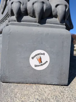 Street sticker A circular white sticker featuring a stylized illustration of a glass of amber beer with a frothy white head at its center. Above the beer, the word 'Stammtisch' is written in a blackletter (Fraktur) style font, curving slightly upwards. Below the beer, the word 'Uttershausen' is written in the same blackletter font, curving slightly downwards. The sticker is applied to a textured, grey, possibly concrete or stone surface.