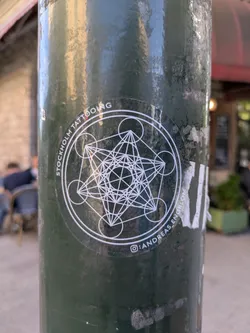 Street sticker The sticker is circular and features a complex geometric design in white on a transparent background, allowing the dark green pole to show through. The central element is a detailed representation of Metatron's Cube, a sacred geometry symbol composed of 13 circles connected by straight lines, creating a three-dimensional cube-like structure. This is enclosed within a larger circle, which in turn is surrounded by another ring. The text 'STOCKHOLM TATTOOING' curves along the top inner edge of the outer ring, and ':ANDREAS.ENGBLOM' along the bottom inner edge, with a small camera icon preceding the latter. The sticker appears to be slightly worn and is attached to a dark green, weathered pole.