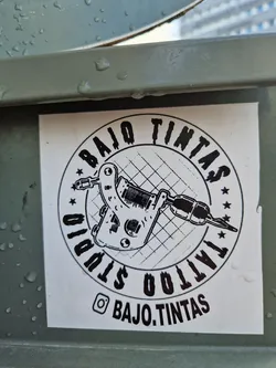 Street sticker Black and white sticker with a tattoo machine illustration and the studio's name.  The sticker is circular with the text 'BAJO TINTAS TATTOO STUDIO' surrounding an image of a tattoo machine. The Instagram handle is also included.