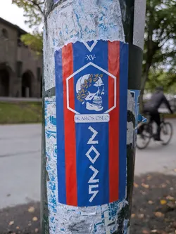 Street sticker A vertical rectangular sticker affixed to a public pole. The sticker features a blue background flanked by two vertical red stripes. At the top, there is a white 'V' shape above the Roman numerals '-XV-'. In the center, a white hexagon contains a white skull in profile wearing a golden-yellow laurel wreath. Below this is a white horizontal bar containing the text 'IKAROS OSLO'. The bottom section features the stylized white numbers '2025' written vertically.