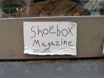 Street sticker A rectangular white paper sticker with a textured surface, stuck to a grey frame. It displays 'Shoebox Magazine' in black handwritten-style lettering, with a printed Instagram handle and website URL at the bottom.