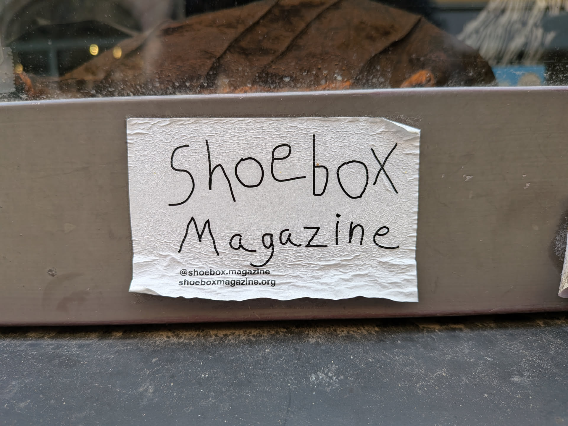 A rectangular white paper sticker with a textured surface, stuck to a grey frame. It displays 'Shoebox Magazine' in black handwritten-style lettering, with a printed Instagram handle and website URL at the bottom.
