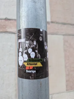 Street sticker A rectangular sticker with rounded corners affixed to a metal pole. The background is a black-and-white photograph depicting a crowd of people wearing blank white masks. Overlaid on the image is text in various colored blocks: 'hurra' in a light pink box, 'för' in a grey box, 'Grönsvarta' on a green banner, 'Attentat' on a yellow banner, 'i' on a red banner, and 'Sverige' on a black banner.