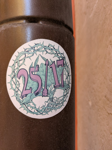 The sticker is circular and features the text "25/17" in purple, with an arrow replacing the slash. The background includes green foliage and a skull. A thorny wreath surrounds the design.