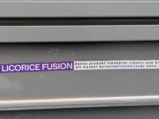 Street sticker The image shows a sticker on a grey corrugated surface. The sticker is horizontally oriented, with the text "LICORICE FUSION" in purple on the left side, followed by a white label with the text "Denna produkt innehåller nikotin som är ett mycket beroendeframkallande ämne."