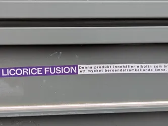 Street sticker The image shows a sticker on a grey corrugated surface. The sticker is horizontally oriented, with the text "LICORICE FUSION" in purple on the left side, followed by a white label with the text "Denna produkt innehåller nikotin som är ett mycket beroendeframkallande ämne."