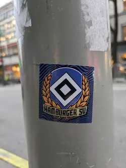 Street sticker A sticker featuring the logo of Hamburger SV, a German football club. The logo is a stylized diamond shape with a wreath. The text "Hamburger SV" is visible at the bottom of the sticker. The sticker has a wavy blue background.