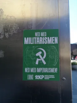 Street sticker A rectangular green sticker featuring bold white text at the top and bottom. The top text reads 'NED MED MILITARISMEN' (Down with Militarism) and the bottom text reads 'NED MED IMPERIALISMEN!' (Down with Imperialism!). In the center, a prominent white hammer and sickle symbol is overlaid on a faint white outline of a globe, depicting continents and grid lines. At the bottom left, a small QR code is present, next to the text 'marxist.se'. To the right of the QR code, the white acronym 'RKP' is displayed, followed by the full name 'REVOLUTIONÄRA KOMMUNISTISKA PARTIET' (Revolutionary Communist Party) in smaller white text.