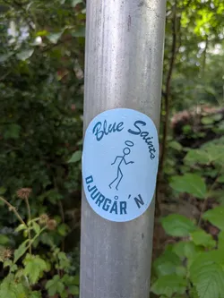 Street sticker The sticker is circular with a light blue background. In the center, a simplistic, black stick figure is depicted in a dynamic pose, possibly skateboarding or dancing, with a small halo above its head. The words "Blue Saints" are written in a stylized font in dark blue above the figure, and "DJURGÅR'N" is written in a similar font below. The sticker appears to be adhered to a metallic pole.