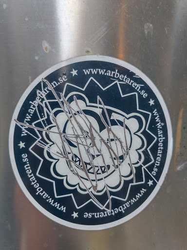 Street sticker A round sticker with the text "www.arbetaren.se" repeated around its perimeter.  A dark blue circular design with a lighter central graphic is present, though heavily defaced with dark scribbles. The sticker appears to be affixed to a metallic surface.