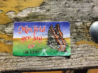 Street sticker A sticker featuring a butterfly with the text "Min frid ger jag er" (Swedish for "My peace I give you") and a bible verse reference Joh, 14:27. The sticker is affixed to weathered wood.