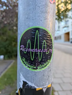 Street sticker The sticker is circular with a vibrant lime green border. The background is black with a textured, almost distressed appearance, scattered with small white speckles. Dominating the center is a stylized, wavy green line resembling a sound wave or oscilloscope reading. Overlaying the lower part of this wave, in a flowing, cursive font, is the word 'Resonansi' in a pinkish-purple hue. Below this, in a smaller, more blocky green font, are the words 'Helsinki is fuckin lit' which appear to be slightly warped to fit the curvature of the sticker.