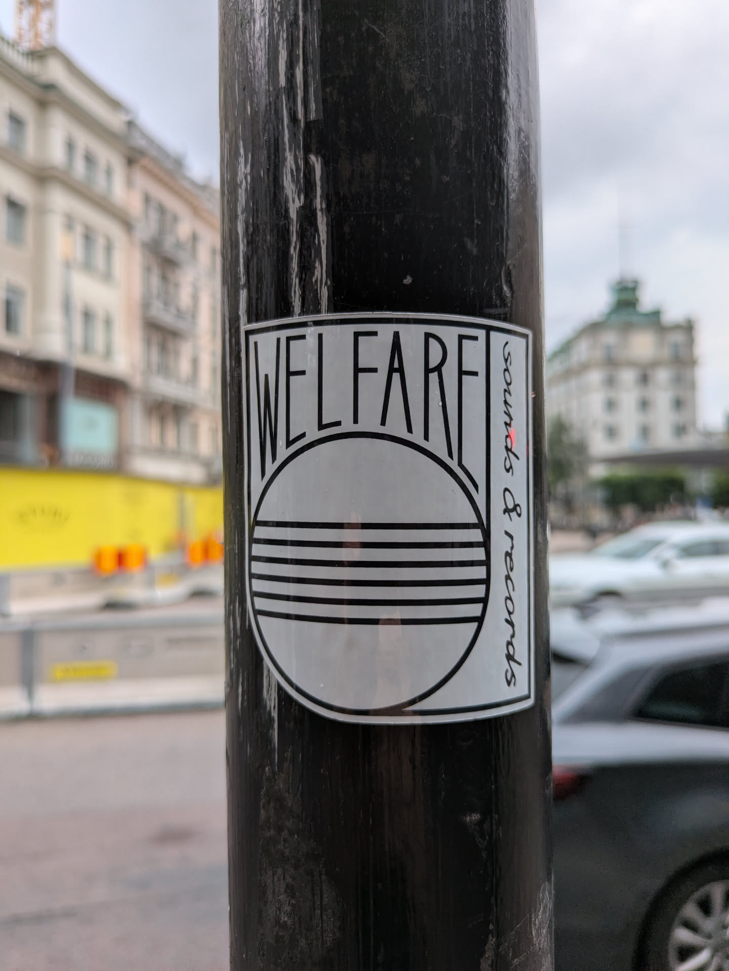 A monochromatic sticker with a rectangular shape and rounded corners, featuring bold black text on a white background. The word "WELFARE" is prominently displayed across the top. Below it, a large circle contains three parallel horizontal lines. Along the right side, oriented vertically, are the words "sounds & records" in a smaller font. The sticker is placed on a dark, weathered pole with visible texture.