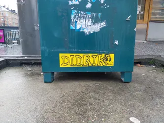 A yellow sticker with the name DIDRIK written in a pixelated font. There is a small drawing to the right of the text. The sticker is placed on a teal colored metal bin outside.