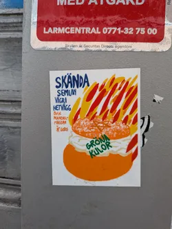 The sticker features a stylized illustration of a semla, a traditional Swedish cream bun, against a vibrant background of red and yellow flame-like streaks. The semla itself is depicted with an orange base, a generous dollop of white cream, and a lighter orange top dusted with white specks. Green text 'GRÖNA KULOR' (Green Balls) is written across the bottom part of the cream. On the left side, blue text reads 'SKÄNDA SEMLAN' (Shame the Semla), followed by 'VÄGRA HETVÄGG' (Refuse Hot Wall), and then orange text 'SVIK MANDELMASSAN' (Betray the Almond Paste) and 'ÄT GODIS' (Eat Candy). The overall style is bold and cartoonish with a clear white border around the central image.