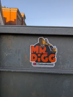 Street sticker The sticker features a bold, orange 'MR. DIGG' text against a dark grey, almost black, irregular circular background with a white outline. An illustration of a yellow and black excavator is positioned above and to the right of the text, with its bucket extended over the 'MR.' part of the text, appearing to be 'digging' into it. Below the main text, there's a smaller white rectangular strip with a red YouTube play button logo and the handle '@TheRealMrDiGG' in black text.
