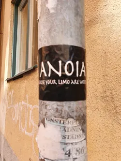 Street sticker A black sticker with white text on it. The text says "ANOIA" in a stylized font, and below it, in a smaller font, it says "Made your limo are wood". The sticker is attached to a pole.