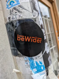 Street sticker The sticker is circular with a matte black background. Centered on it is the word 'beWider' in a vibrant orange, sans-serif font. The 'b' features a short horizontal line extending from its top-left curve, and the 'r' has a similar horizontal line extending from its top-right curve, giving the text a distinctive, stylized appearance. The sticker stands out against the worn and layered stickers on the pole.