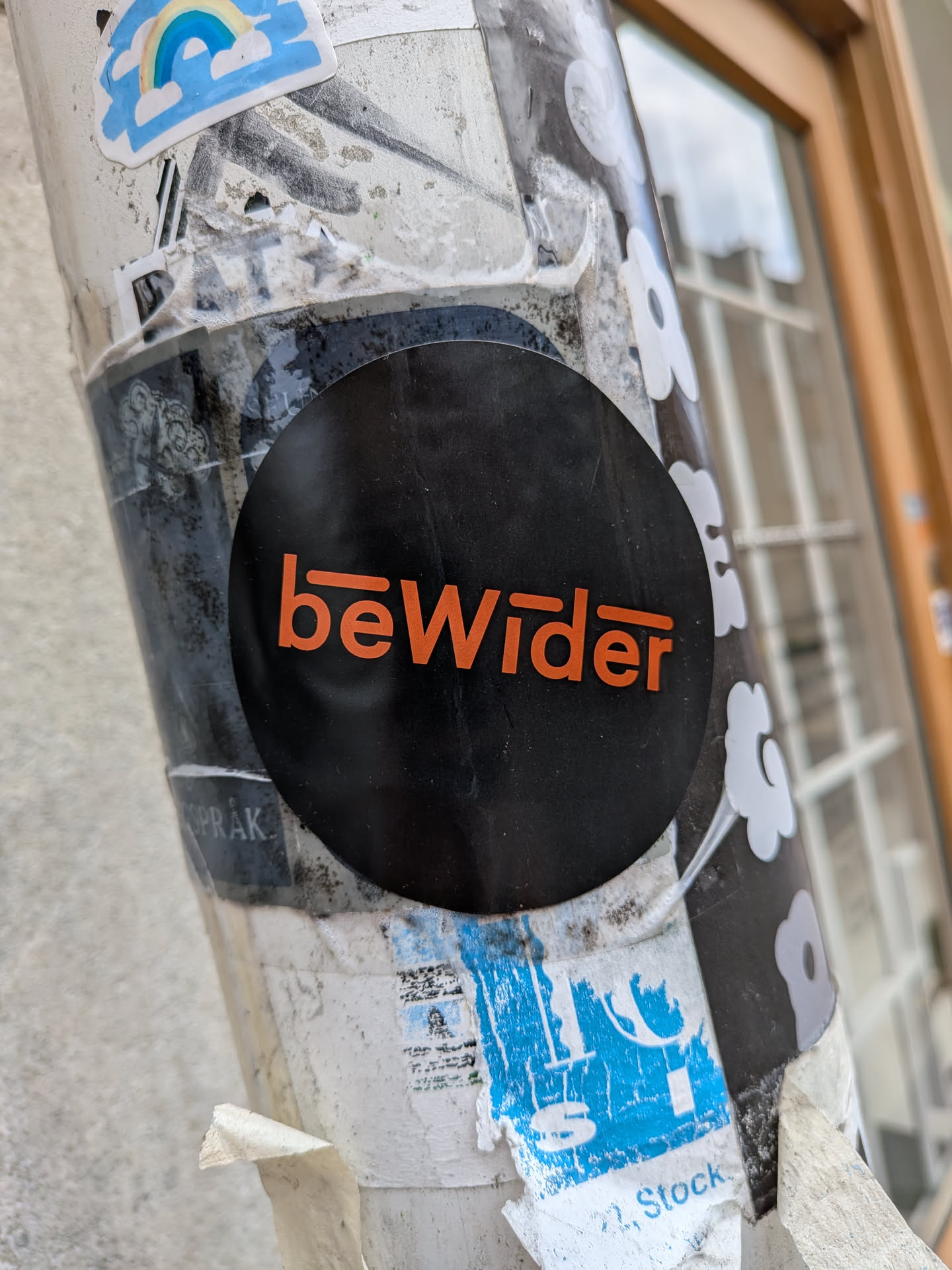 The sticker is circular with a matte black background. Centered on it is the word 'beWider' in a vibrant orange, sans-serif font. The 'b' features a short horizontal line extending from its top-left curve, and the 'r' has a similar horizontal line extending from its top-right curve, giving the text a distinctive, stylized appearance. The sticker stands out against the worn and layered stickers on the pole.