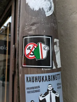 A square sticker featuring a crossed-out hammer and sickle symbol combined with a Finnish political party logo. The sticker is affixed to a pole.