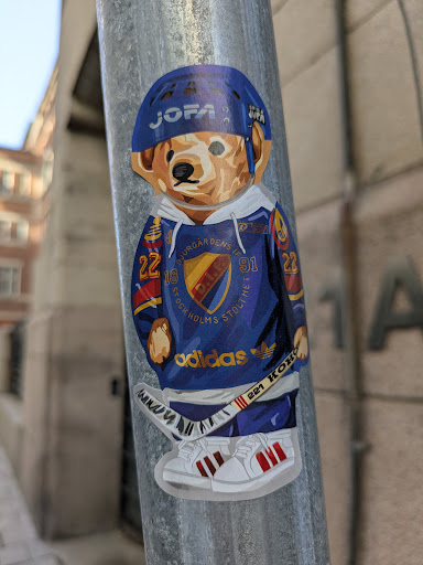 Street sticker The sticker depicts a teddy bear dressed in a hockey jersey, helmet, and sneakers. The bear is wearing a blue JOFA helmet, a blue and yellow Djurgårdens IF hockey jersey with the numbers 22 and 91, and Adidas sneakers. The jersey also features an Adidas logo. The bear is holding a hockey stick with "221 KOHO" written on it. The bear is standing on a metal pole, likely a light post or similar street fixture.