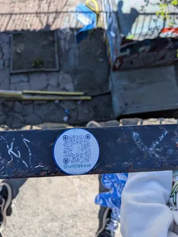 Street sticker A circular white sticker with a thin blue border, affixed to a dark, rusty metal beam. The center of the sticker features a prominent black and white QR code. Above the QR code, in small black text, it reads 'Say hi!'. Below the QR code, there's an Instagram logo followed by '@unclekase' in blue text, and further below, 'linktr.ee/unclekase' in smaller black text.