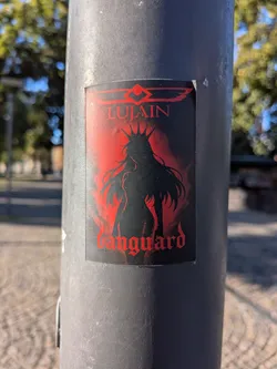 Street sticker The sticker features a predominantly black and red color scheme. At the top, a stylized winged logo is present in red. Below this, the word "LUJAIN" is written in bold, red, gothic-style lettering. The central element is a silhouette of a figure with long, flowing hair, wearing a spiky crown, and a form-fitting outfit. This silhouette is stark black against a fiery red background that fades into black at the edges. Below the figure, the word "vanguard" is also displayed in bold, red, gothic-style lettering. The overall aesthetic is dark and powerful.
