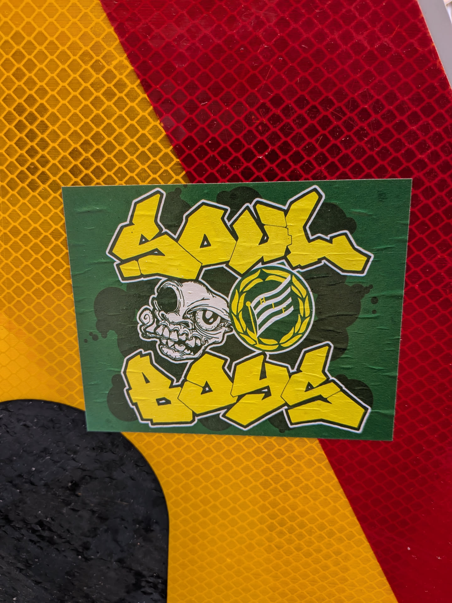A rectangular sticker with a dark green background, subtly textured with darker splatters or shadows. The main design features the words "SOUL" at the top and "BOYS" at the bottom, rendered in bold, yellow bubble letters with a white outline and a black drop shadow. Centered between the words is a cartoonish, grotesque skull-like creature in white with black outlines and details, characterized by a single large eye, prominent teeth, and a distorted facial structure. To the right of the skull is a circular emblem with a yellow laurel wreath border. Inside the emblem, on a dark green background, is a white flag with horizontal stripes, depicted on a pole. There appears to be small, indistinct text on the flag. The sticker itself shows visible creases and slight wear, indicating it's a physical object applied to a surface.