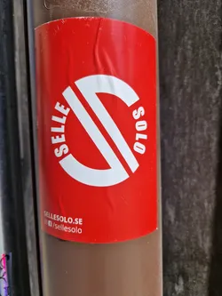 A red and white SelleSolo sticker with a logo is attached to a brown pole. The sticker includes the website sellesolo.se and social media link /sellesolo.
