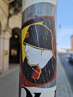 This sticker shows a stylized illustration of a soldier's profile wearing a helmet with a red and yellow emblem on it. The sticker is worn and shows signs of age and weathering, adding to its character. The imagery suggests possible commentary on war, military history, or political ideology, requiring further research to provide more context.