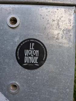 Street sticker A round black sticker with white text reading 'LE VIOLON DINGUE' in a stylized font. Features crossed wheat stalks at the bottom and contact info, including address, phone, website, and Instagram handle, printed along the circular border.