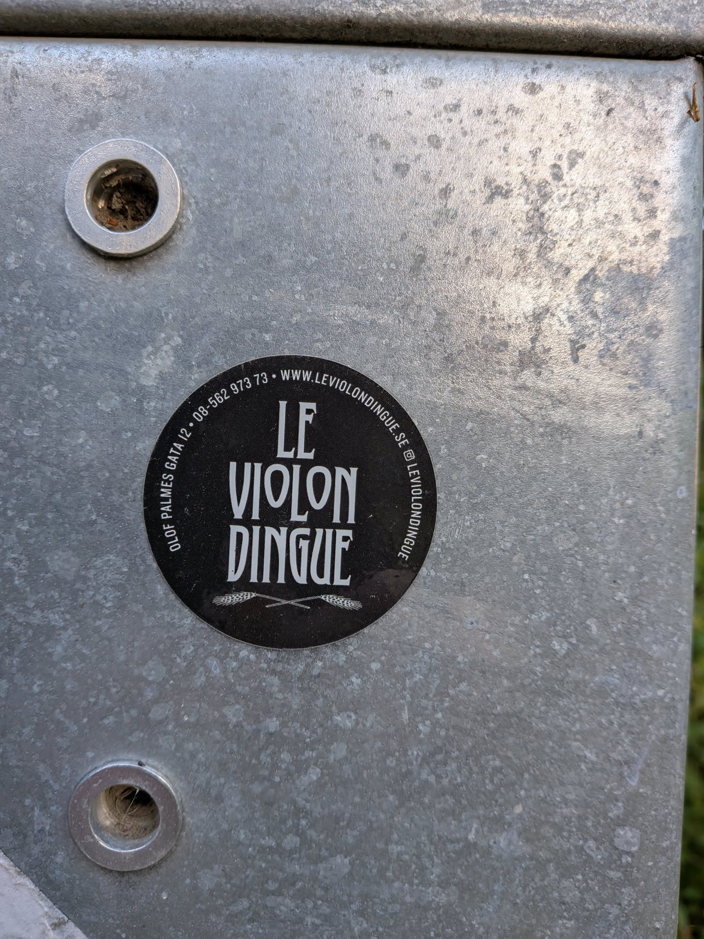 A round black sticker with white text reading 'LE VIOLON DINGUE' in a stylized font. Features crossed wheat stalks at the bottom and contact info, including address, phone, website, and Instagram handle, printed along the circular border.