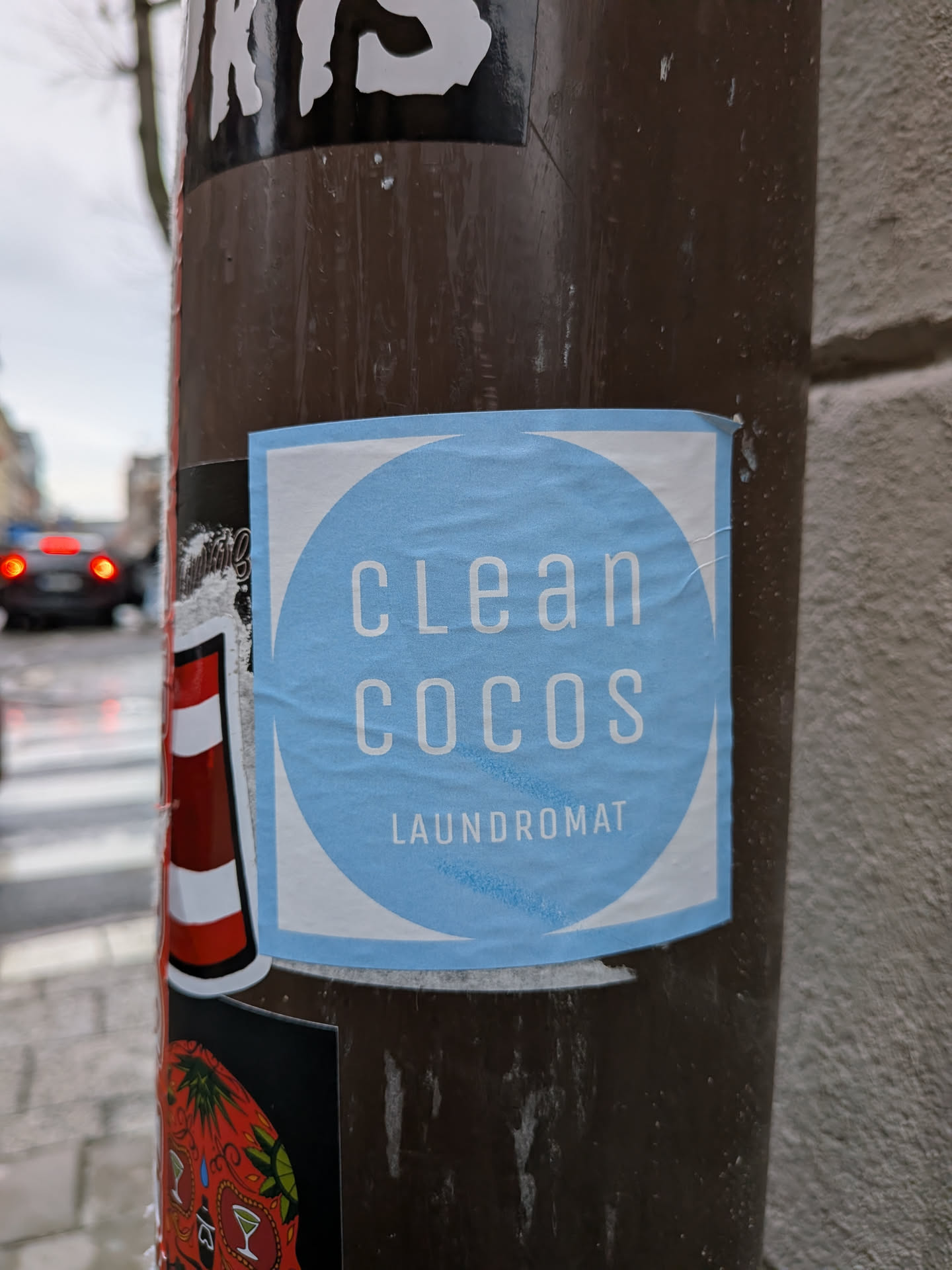 The sticker is square-shaped with a prominent light blue circle in its center, set against a white background. Inside the light blue circle, the words "clean cocos" are displayed in a clean, sans-serif, all-lowercase white font. Below this, in a smaller, all-caps white font, is the word "LAUNDROMAT". The sticker shows signs of being a physical sticker, with slight wrinkles and adherence to a dark brown pole.