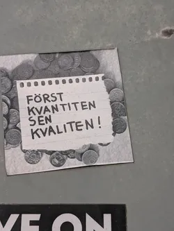 Street sticker A square grayscale sticker featuring a background of scattered coins. In the center, a graphic of a torn piece of notebook paper displays the handwritten Swedish phrase 'FÖRST KVANTITEN SEN KVALITEN!'.