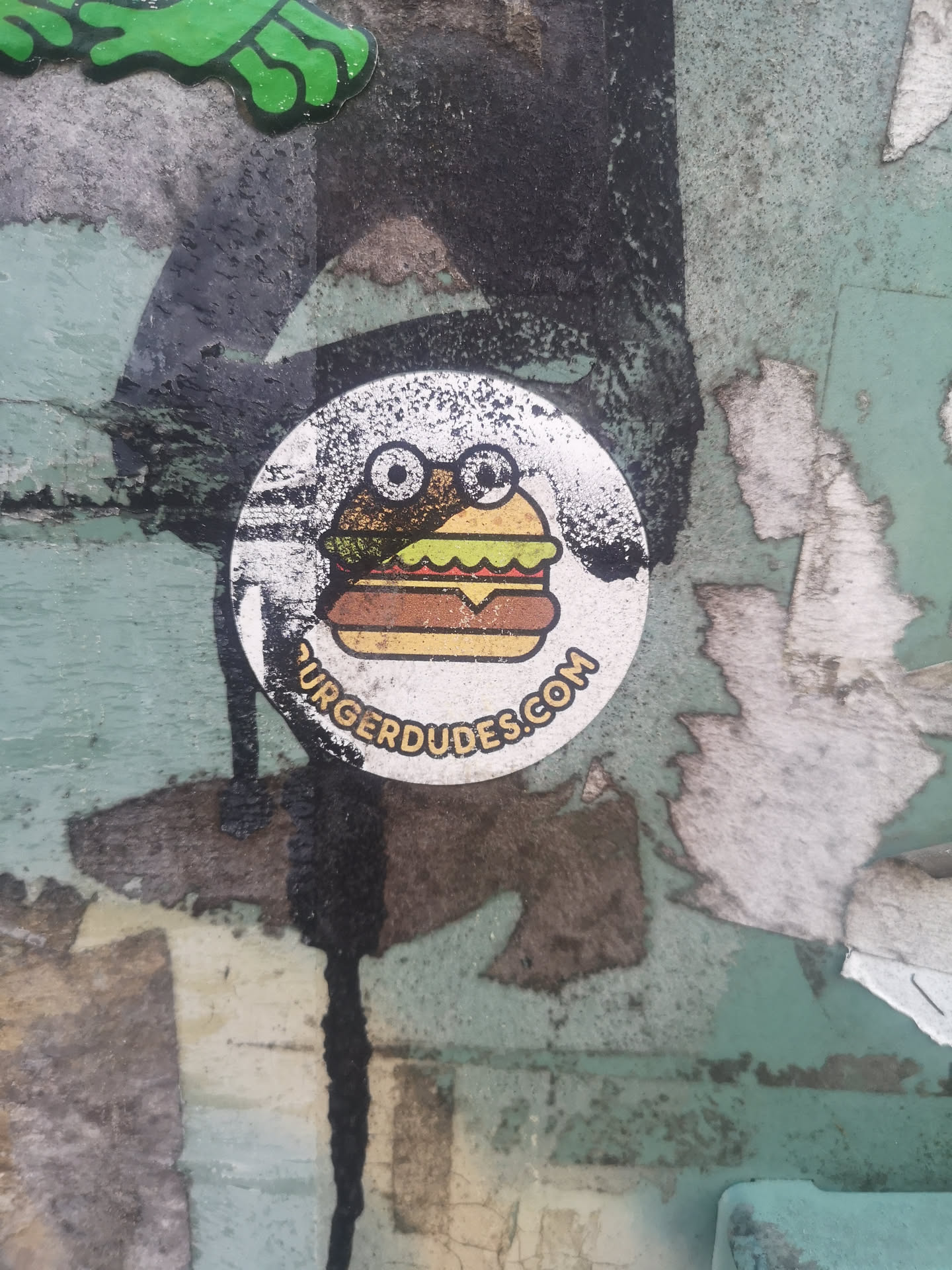 A circular sticker featuring a cartoon-style burger with two prominent, white, googly-like eyes on its top bun, giving it a frog-like or whimsical character. The burger consists of a light yellow bun, a wavy green lettuce layer, a brown patty, and a yellow cheese slice, all outlined in a darker brown. The background of the sticker is primarily white but shows significant wear, with black and grey speckles and distressed areas. Along the bottom curve of the sticker, there is text in an arc.