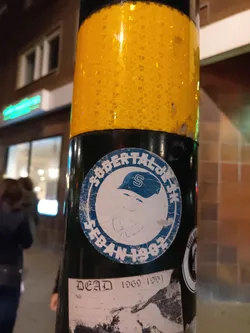 Street sticker A worn sticker featuring a cartoon character wearing a baseball cap with the letter 'S'. The text 'Seban 1982' is visible within a circular design. Below this sticker, there is another sticker with 'Dead 1969-1991' written on it. 