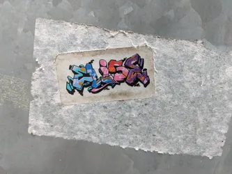 Street sticker The sticker features graffiti-style lettering that appears to spell out the name 'ALON'. The letters are stylized with thick black outlines and filled with a gradient of colors. The first letter is predominantly blue with light blue and pink accents. The second letter is bright pink with orange highlights. The third letter is a mix of orange and pink with darker pink accents. The final letter is a deep purple with orange and yellow accents within its structure, and it has a shadow effect. The sticker itself is slightly aged and worn, with visible texture and some discoloration on its white background, attached to a rough, textured grey surface.