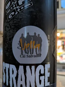 Street sticker The sticker is circular with a light grey background and a thin, dark border. In the center, there are four dark grey silhouettes of people standing closely together, with one person's arm around another's shoulder. Above the silhouettes, the word 'Colla' is written in a stylized orange cursive font. Below the silhouettes, the words 'Cal Salvador' are written in a simple black sans-serif font. The sticker appears to be slightly aged.