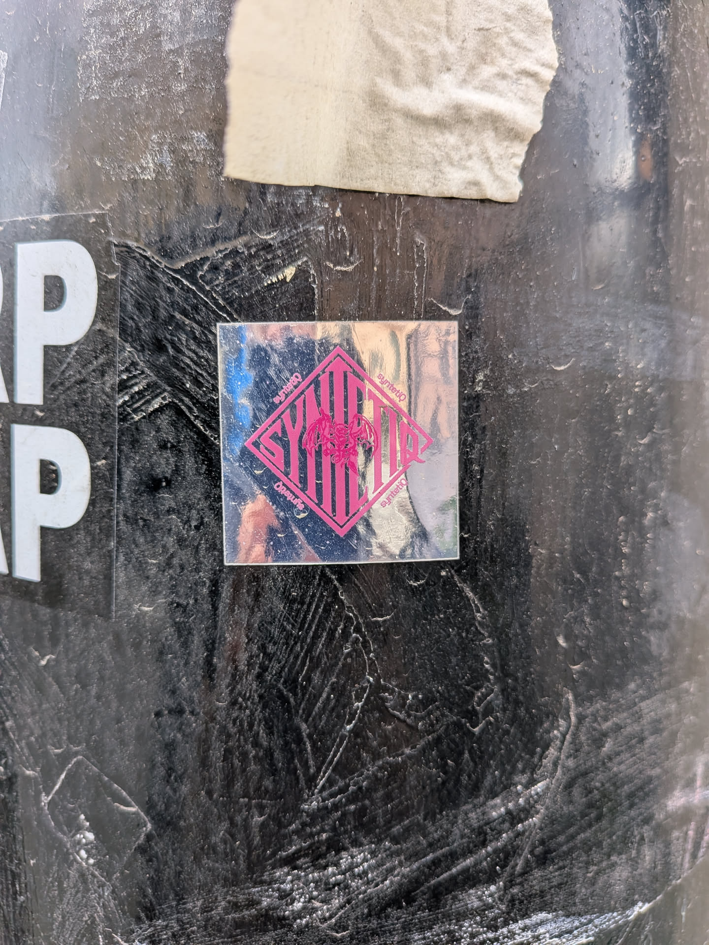 A square, holographic sticker featuring a bold design in vibrant pink. The central element is a large diamond shape, filled with vertical stripes and the stylized, vertically oriented text 'SYNTETIQ'. Overlaid on the text and within the diamond is a detailed illustration of a winged, dragon-like creature with sharp claws and teeth. Smaller, curved text reading 'syntetiq' appears along the top and bottom edges of the diamond. The holographic material reflects various colors, including blues, purples, and silvers, creating a shimmering effect against the dark, textured surface it's adhered to.