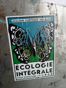 Street sticker The sticker is a rectangular piece of graphic art, predominantly featuring a black butterfly with intricate white, blue, and green patterns on its wings, set against a split background of teal on top and green on the bottom. The upper wings display abstract organic shapes, including what appears to be a fetus-like figure and other natural elements. The lower wings contain symbols such as an atom and flowers, suggesting a blend of scientific and natural themes. Above the butterfly, the text "LOCALISME • BIOÉTHIQUE • NUCLÉAIRE" is prominently displayed. Below the butterfly, in large bold letters, it reads "ÉCOLOGIE INTÉGRALE". At the very bottom, the words "LYON POPULAIRE" are visible, alongside icons indicating social media presence on Facebook, X (formerly Twitter), Instagram, and Telegram.