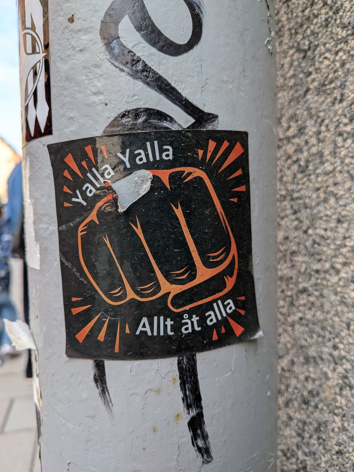 A square-shaped sticker with a black background, featuring a prominent, stylized orange outline of a clenched fist at its center. Orange, triangular rays emanate outwards from the fist, suggesting energy or impact. White text 'Yalla Yalla' is curved above the fist, and 'Allt åt alla' is curved below it. The sticker shows signs of wear, including a small tear near the top of the fist, and is affixed to a light grey, painted pole.